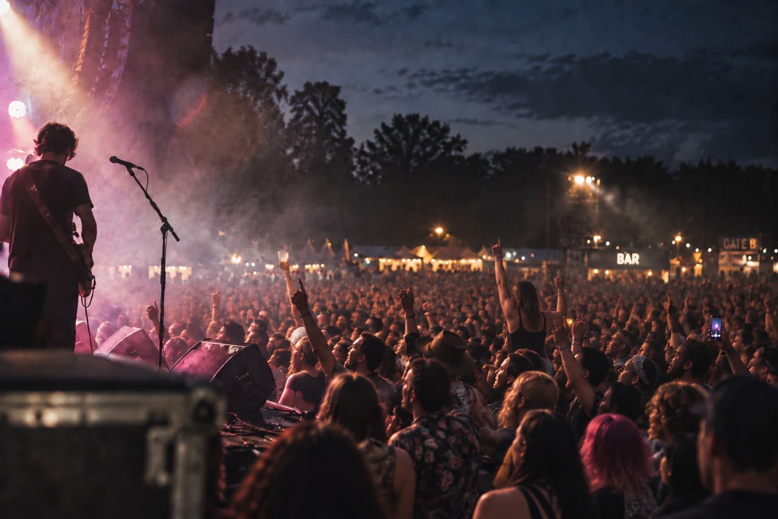 A festival crowd at dusk seen from the side of the stage, a guitarist in the foreground