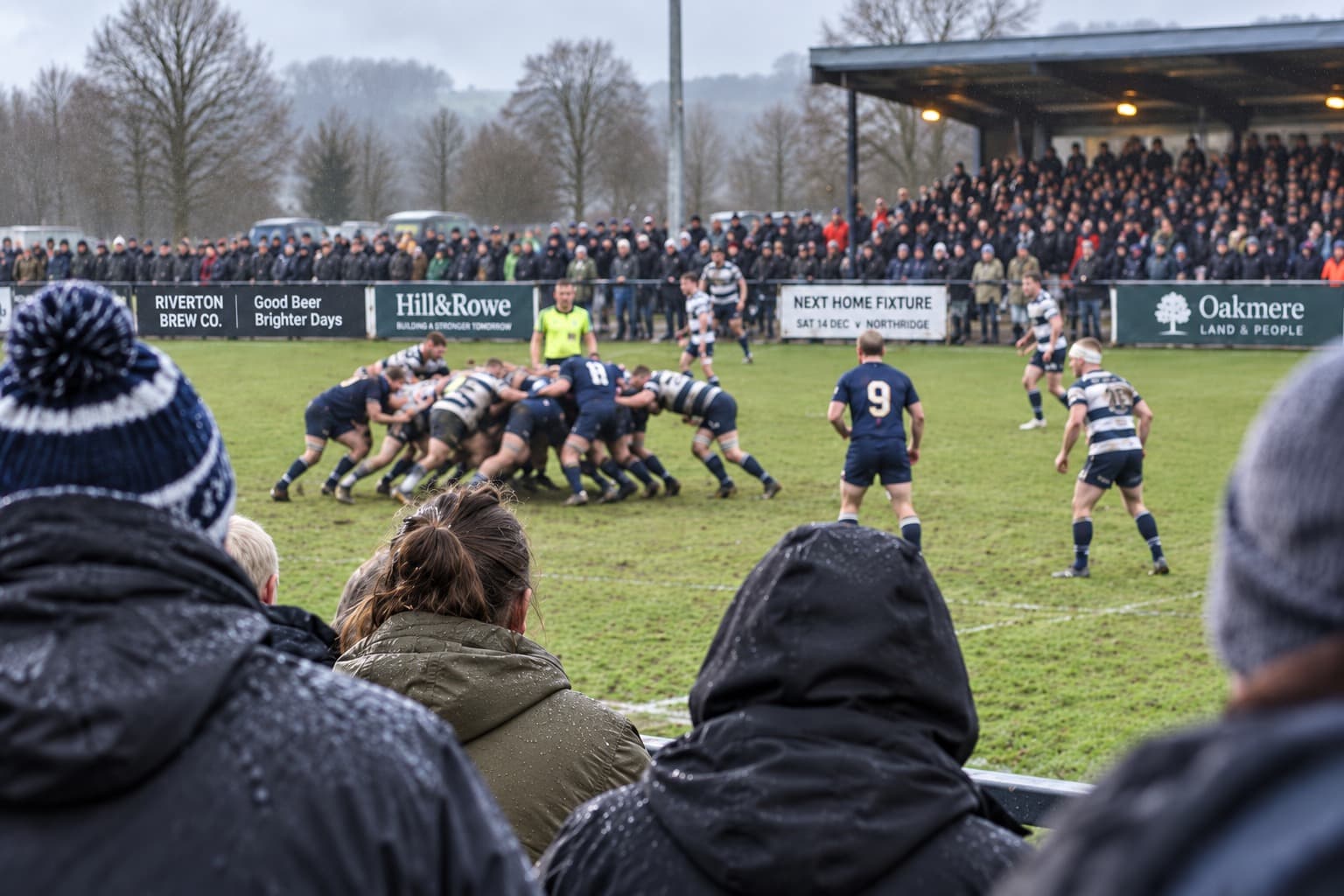 A club rugby scrum on a wet afternoon, seen over the shoulders of supporters at the rail
